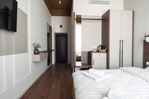 A hotel room with a wooden floor, a bed with white bedding, a TV mounted on the wall, a wardrobe, and a small table with a vase.