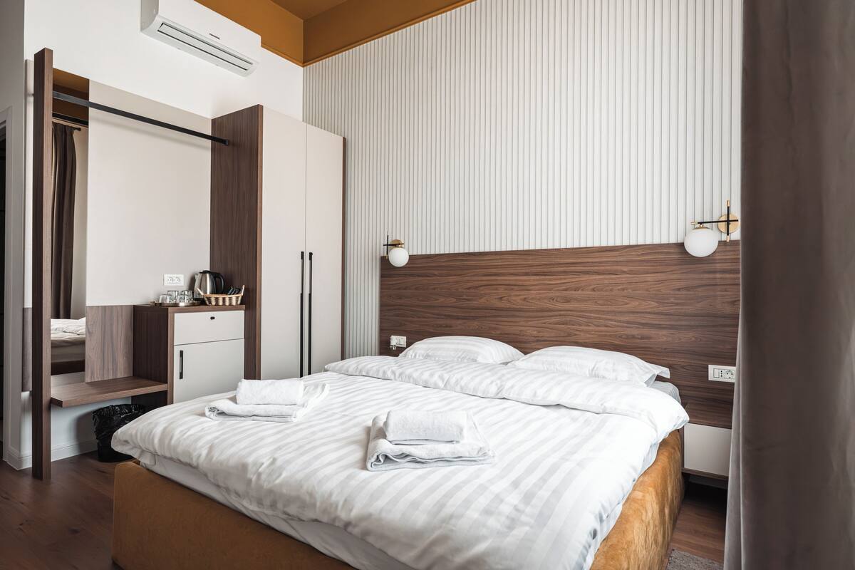 A modern hotel room with a wooden headboard, a large bed with white bedding, a bedside table with a lamp, and a wardrobe.