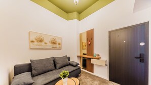 A modern living room with a grey sofa, a small round table with a plant, a painting on the wall, a wooden cabinet, and a dark door.