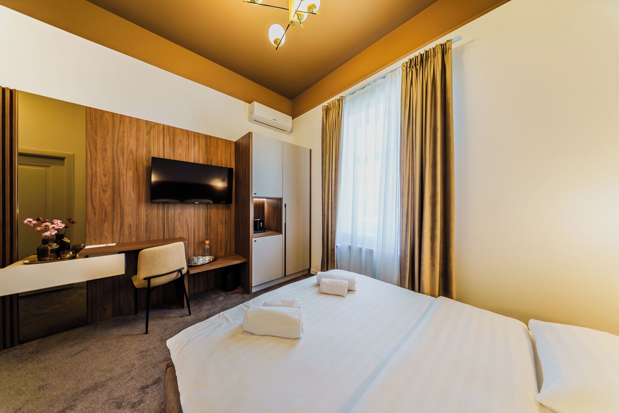 Quarto de hotel com cama, escrivaninha, cadeira, televisão e janela com cortinas.