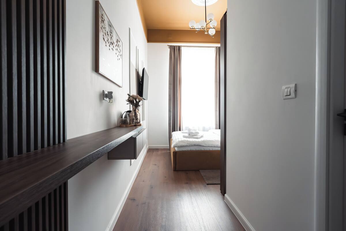 A hallway with a wooden floor, a bed with white bedding, a mounted TV, and a ceiling light fixture.