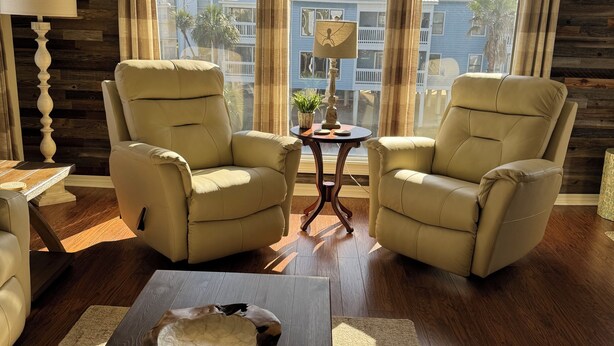 A living room with two beige armchairs, a wooden coffee table, a ceiling fan, and a view of buildings through large windows. - Spacious 2-bedroom condo at Sea Oats -D206 in beautiful Gulf Shores, Alabama (Gulf Shores)