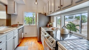 Una cocina con armarios blancos, un refrigerador de acero inoxidable, una estufa y una ventana con vista a palmeras.