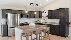 Une cuisine moderne avec des meubles de couleur sombre, des appareils en acier inoxydable, un îlot central en bois avec des tabourets de bar, un éclairage suspendu et un pare-feu en carreaux.