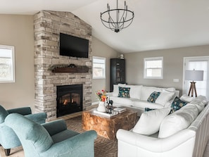 Un salón con chimenea de piedra, televisor de pantalla plana, sofá blanco, sillones color turquesa y una mesa de centro con objetos decorativos.