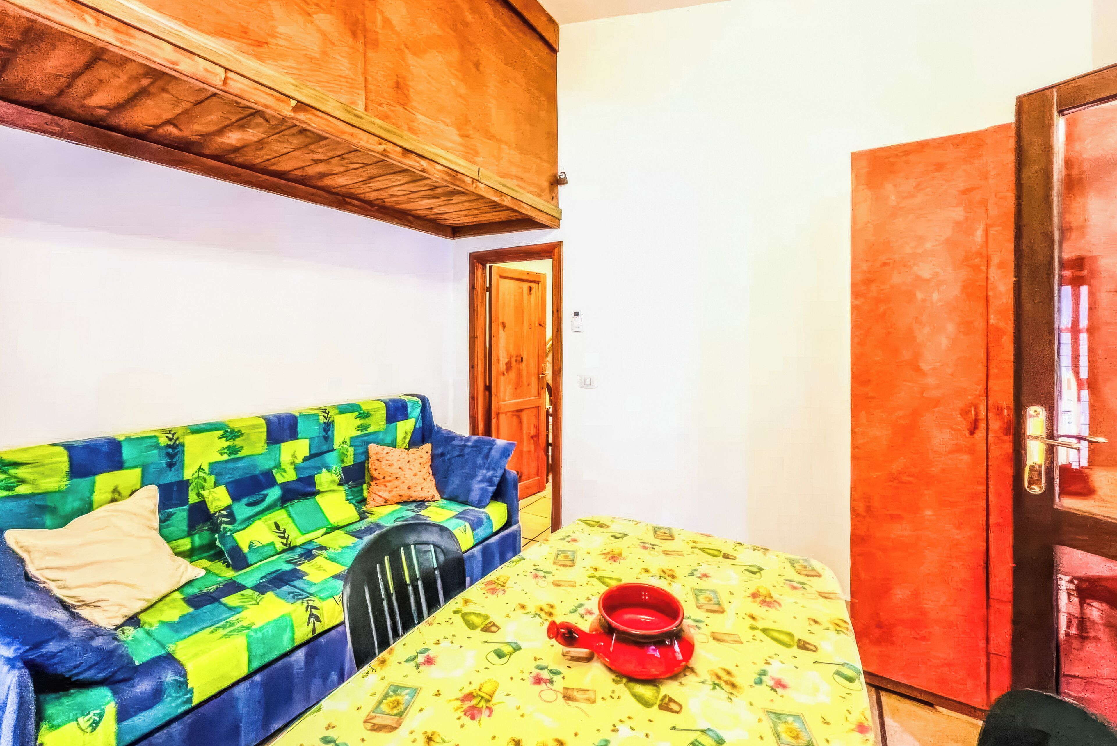 Une pièce avec un plafond en bois, un canapé aux couleurs vives, une table recouverte d’une nappe à motifs floraux et une théière rouge.