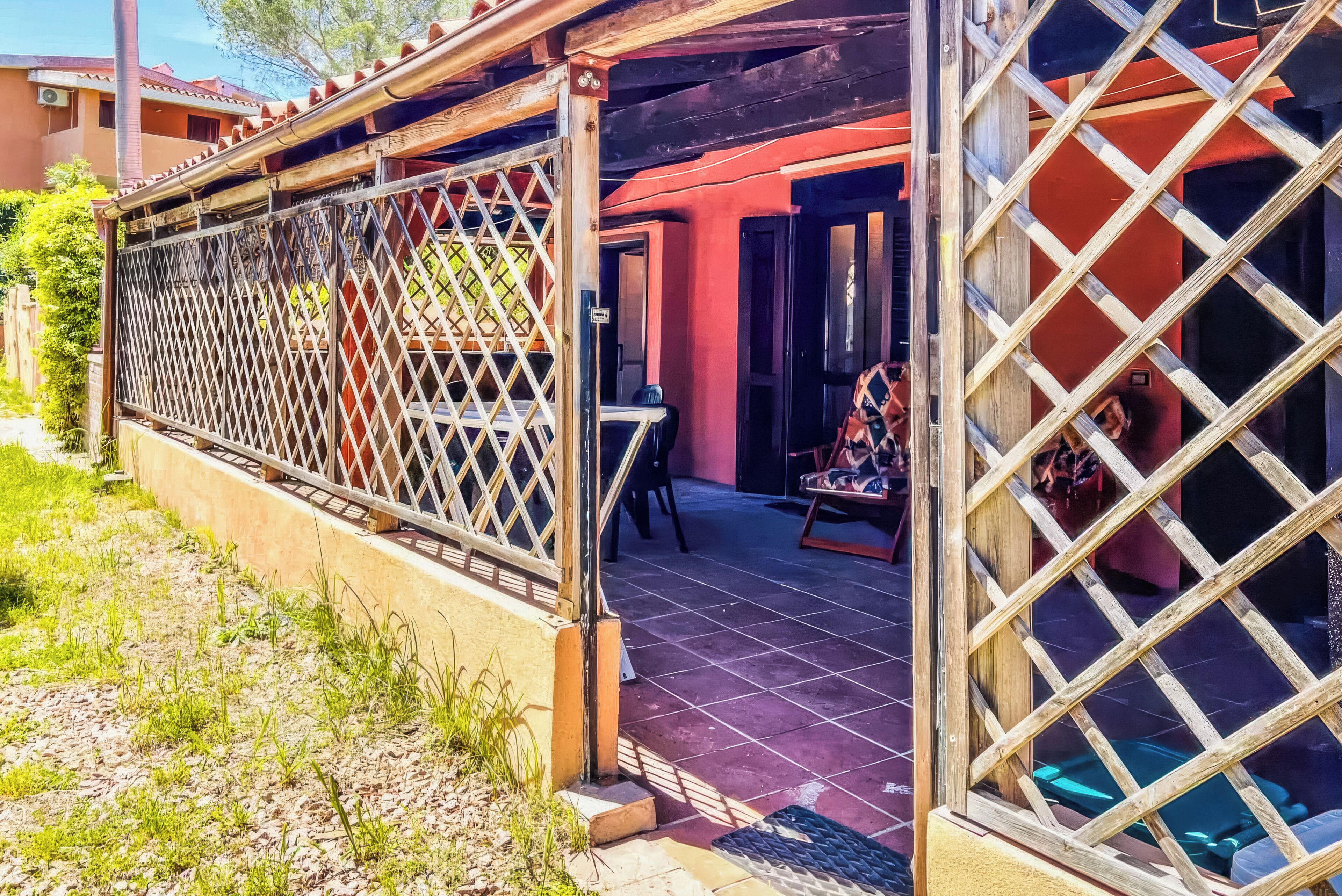 A covered outdoor area with a wooden lattice fence, a tiled floor, and a view of a building with red walls and a door.