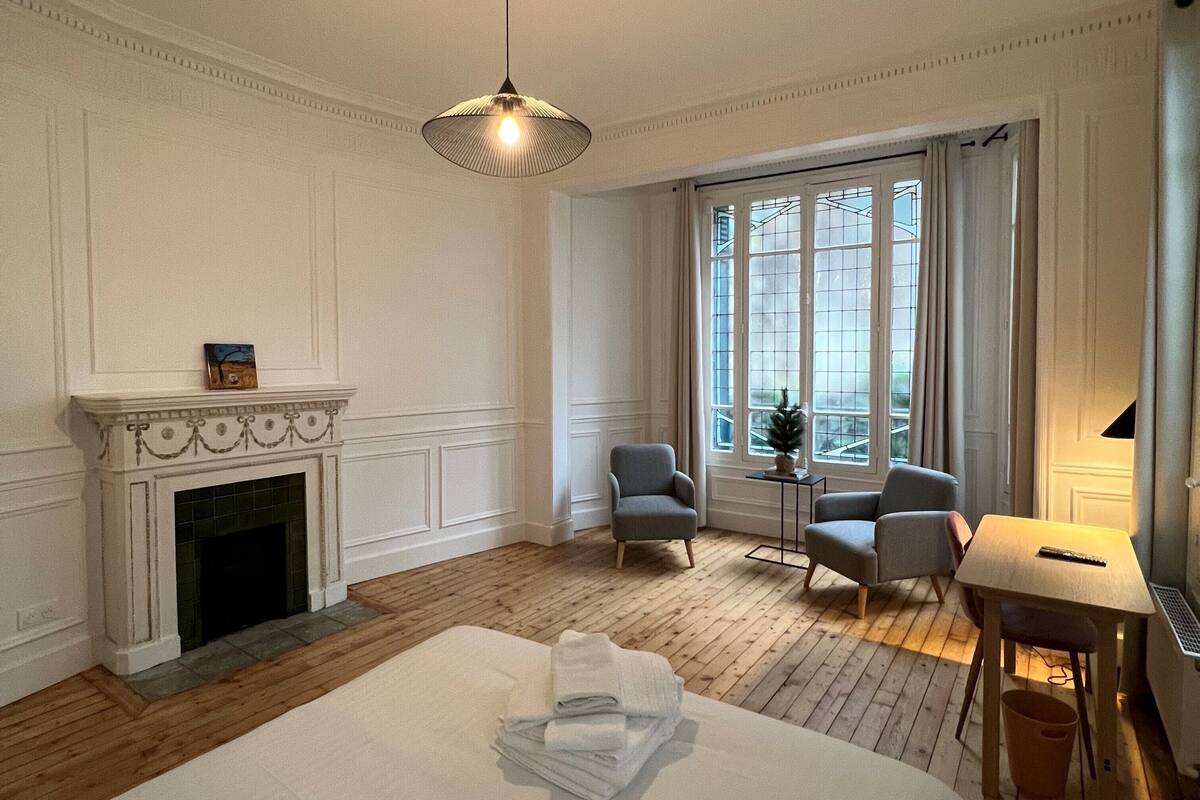 A hotel room with a fireplace, a bed, two armchairs, a small table, and a large window.