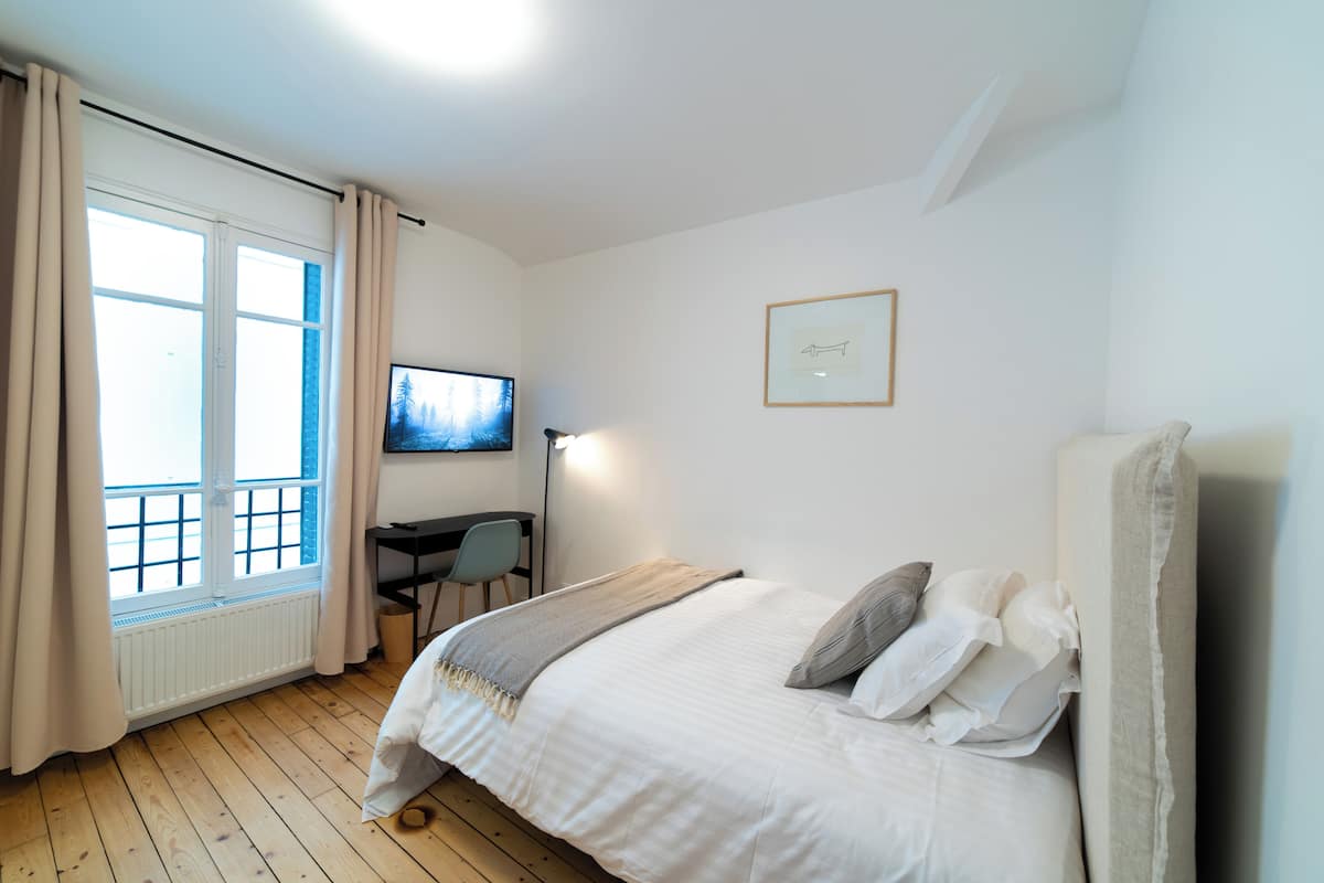 A bedroom with a bed, a desk, a chair, a television, and a window with curtains.