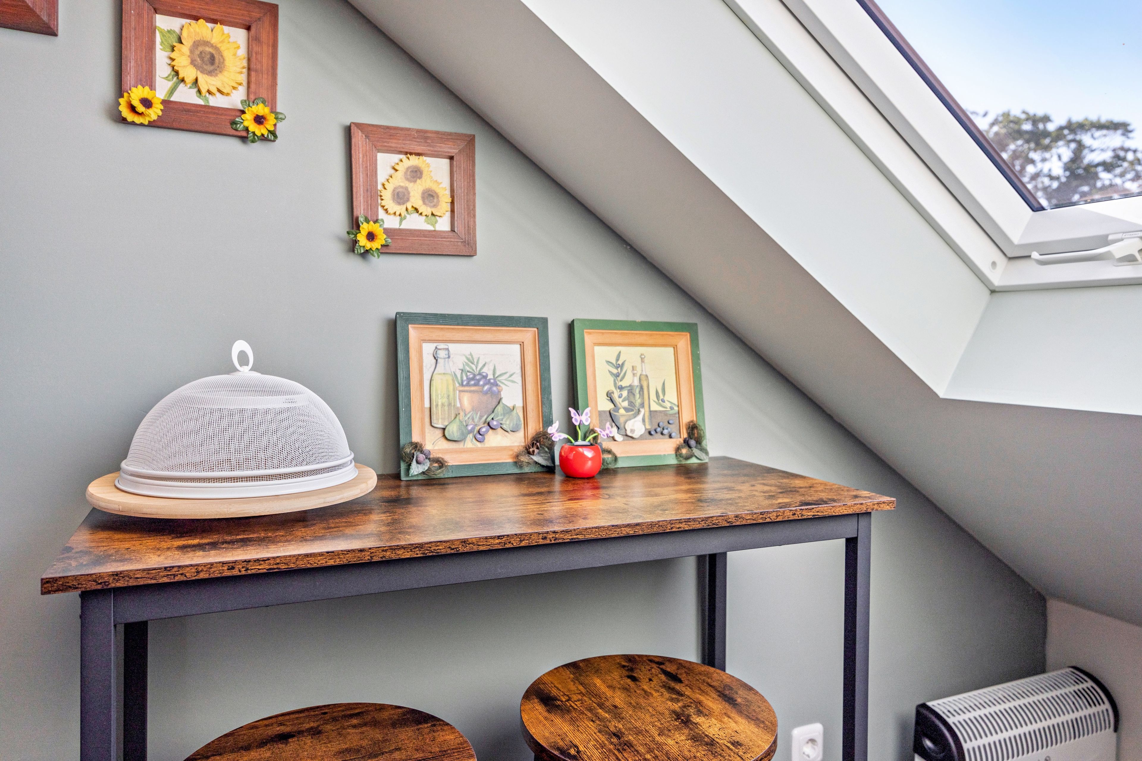 Une pièce avec un plafond en pente, une table sur laquelle se trouve un objet blanc, et deux tableaux encadrés représentant des tournesols.