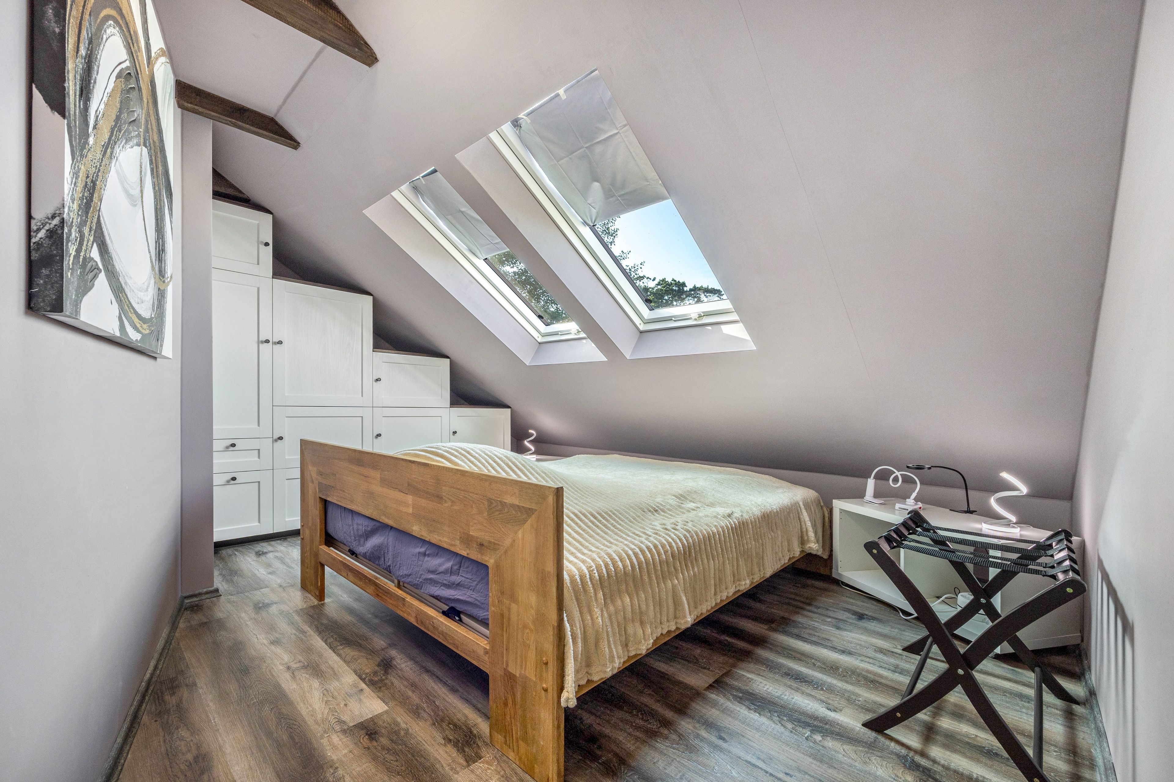 Un dormitorio con techo inclinado, cama, escritorio y silla. Hay tragaluces que permiten entrada de luz natural.