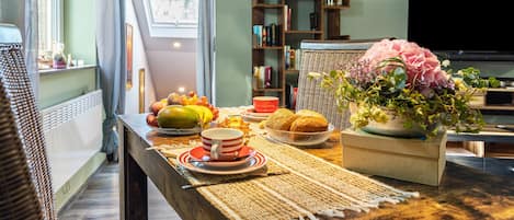 A dining area with a table set for two, featuring a variety of fruits, bread, and a floral centerpiece.