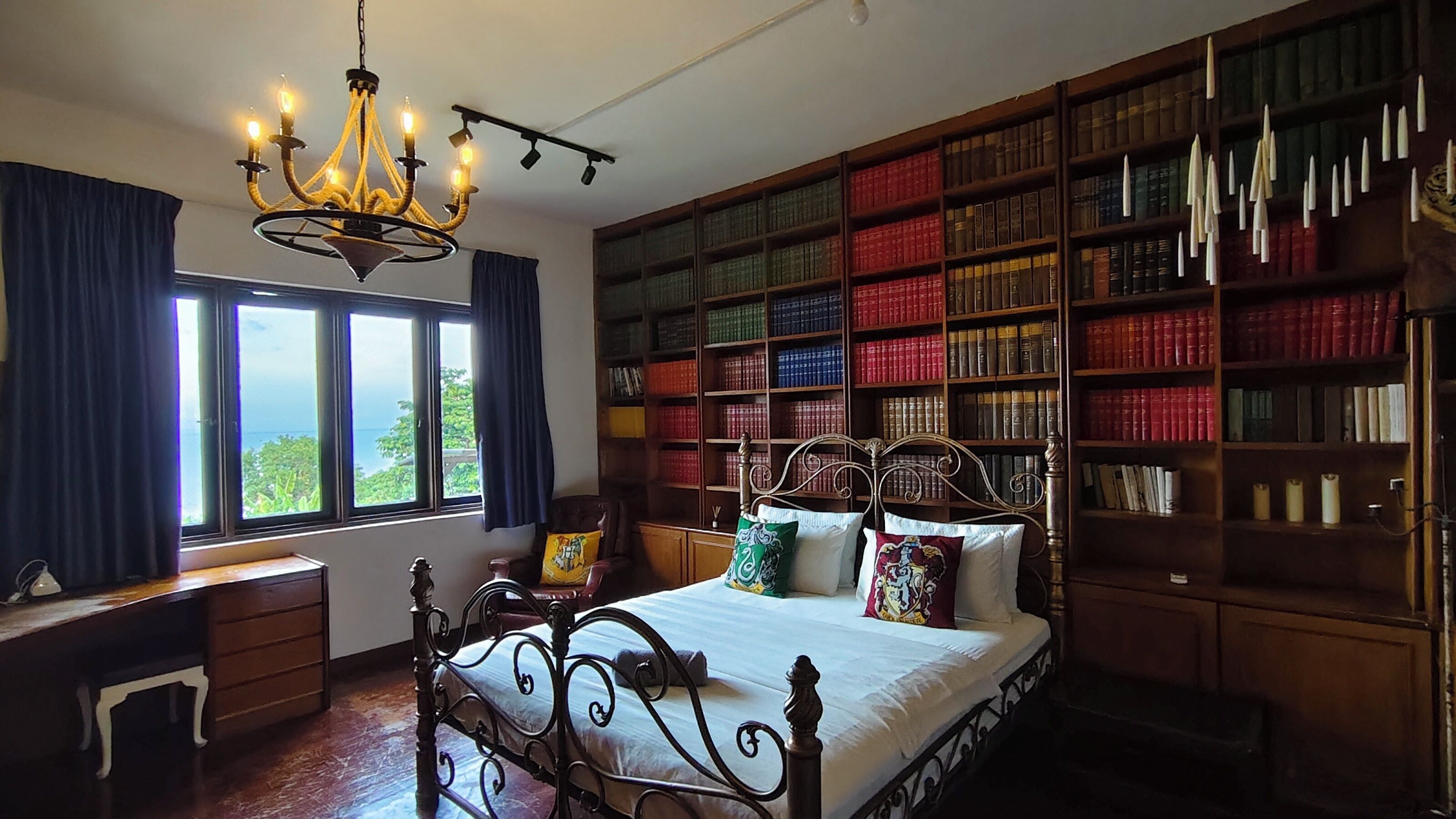 A bedroom with a large bookshelf, a bed with decorative pillows, a desk, and a chandelier.