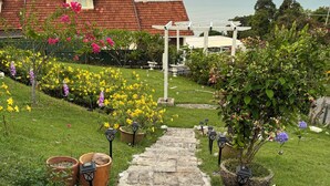 Un sendero en un jardín conduce a una casa con techo de tejas rojas, rodeada de diversas plantas en flor y luminarias exteriores.