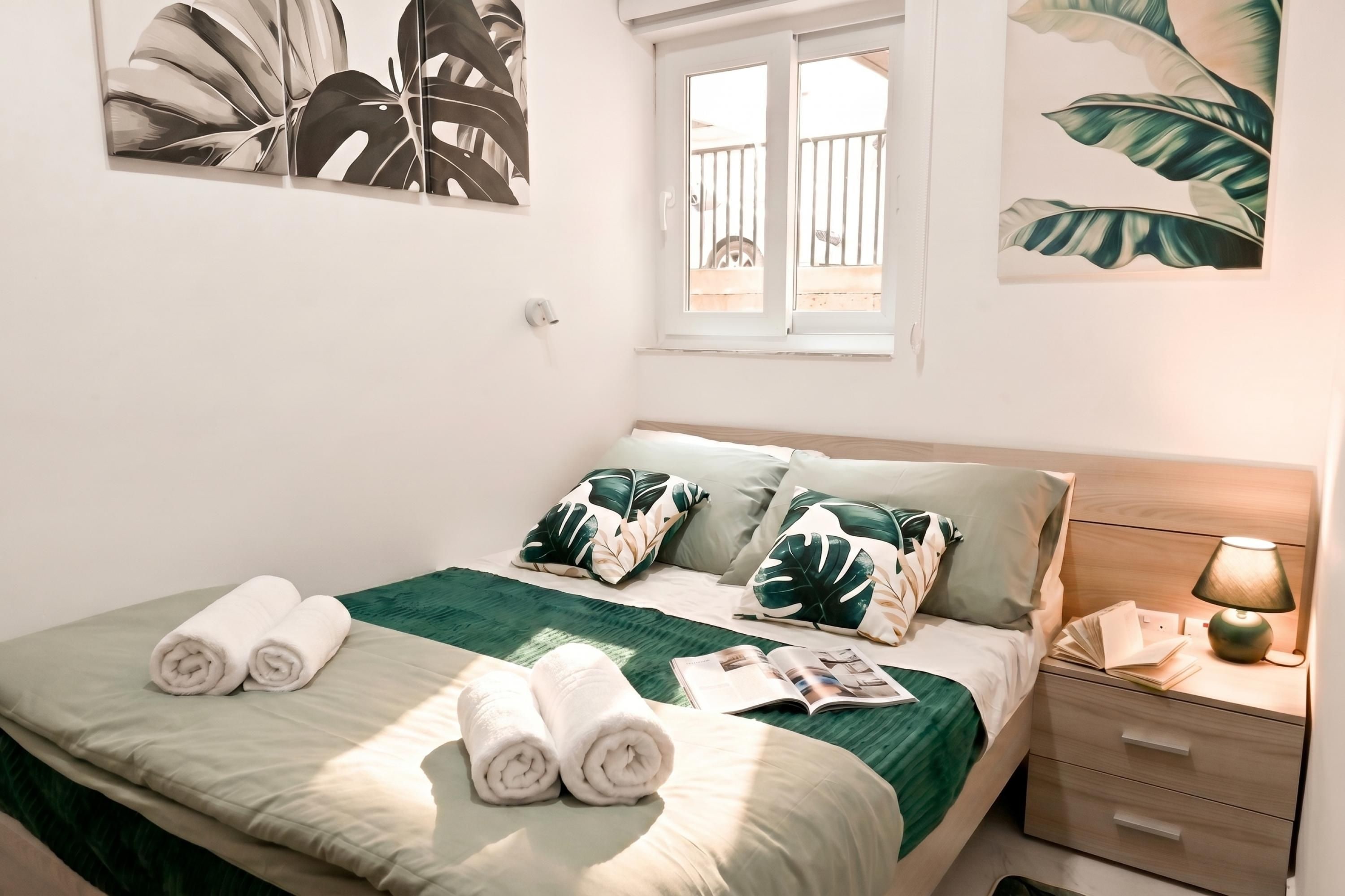Una cama bien hecha con ropa de cama verde y blanca, una ventana con vistas y una mesita de noche con una lámpara.