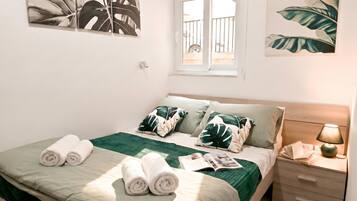 Una cama bien hecha con ropa de cama verde y blanca, una ventana con vistas y una mesita de noche con una lámpara.