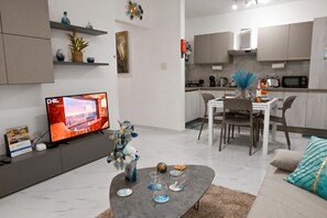 A modern living room with a flat-screen TV, a dining area with a white table and chairs, and a kitchen with grey cabinets and appliances. - Cozy 2bdr Apart In Bugibba Square (St. Paul's Bay)