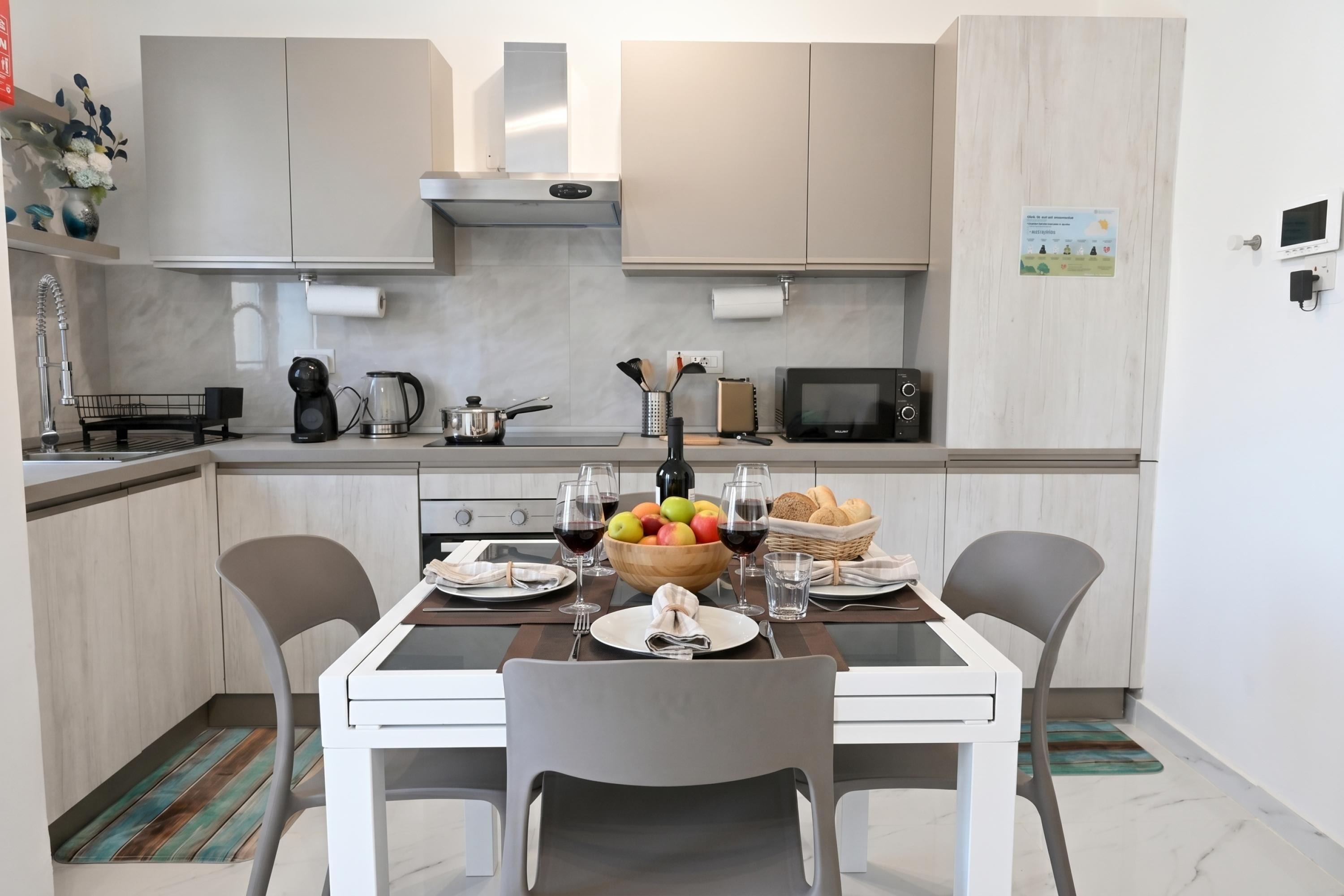Una cocina moderna con una mesa de comedor dispuesta para dos personas, que incluye un frutero, copas de vino y una cesta de pan.