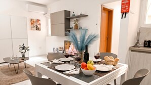 A dining area with a white table, grey chairs, and a bowl of fruit. - Cosy 2bdr Apart In Bugibba Square (St. Paul's Bay)