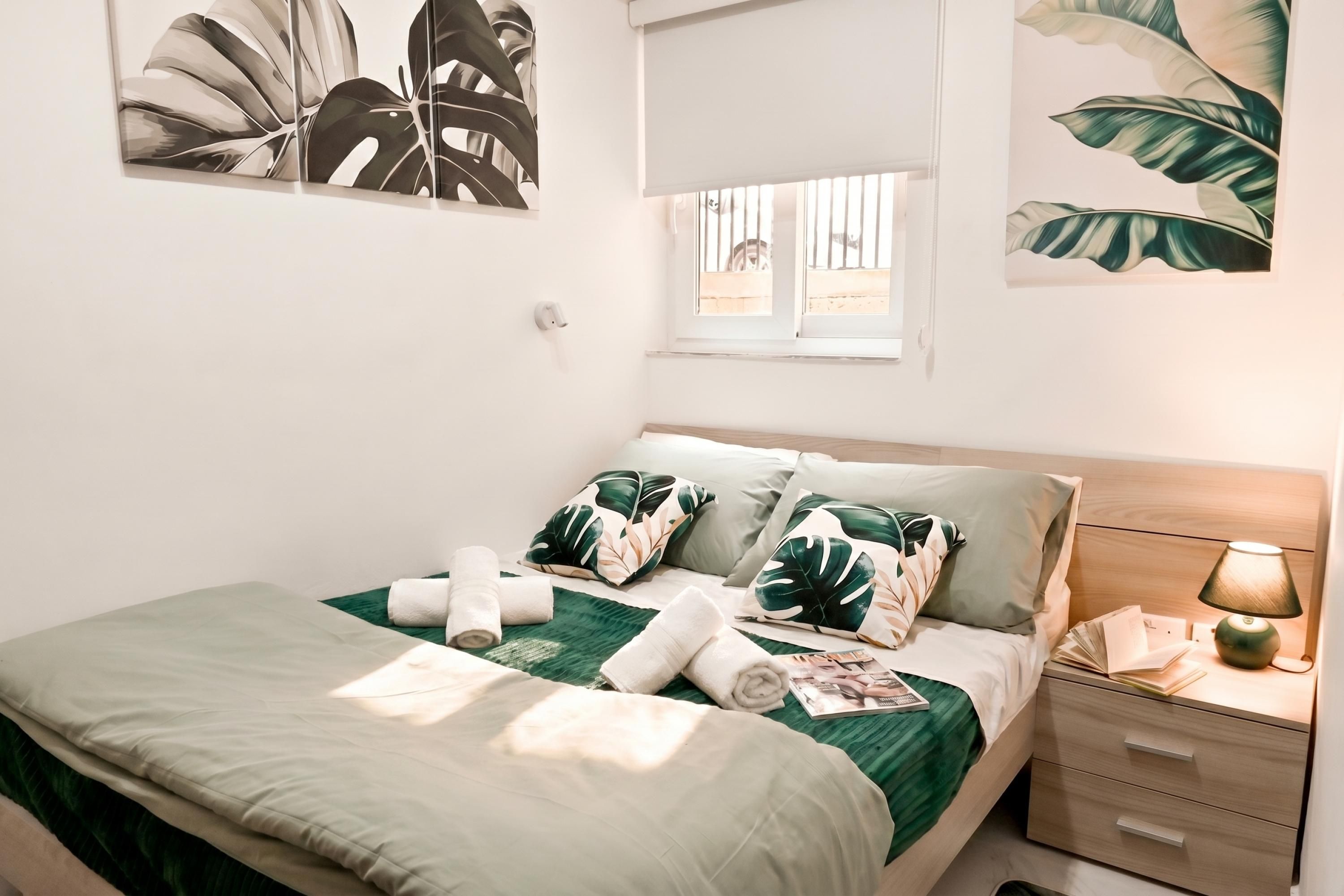 Una cama bien hecha con una colcha verde, almohadas blancas y toallas enrolladas. Hay una mesita de noche con una lámpara y una ventana con barrotes.