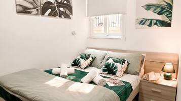 Una cama bien hecha con una colcha verde, almohadas blancas y toallas enrolladas. Hay una mesita de noche con una lámpara y una ventana con barrotes.