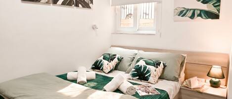 Een net opgemaakt bed met een groene plaid, witte kussens en opgerolde handdoeken. Er staat een nachtkastje met een lamp en een raam met tralies.