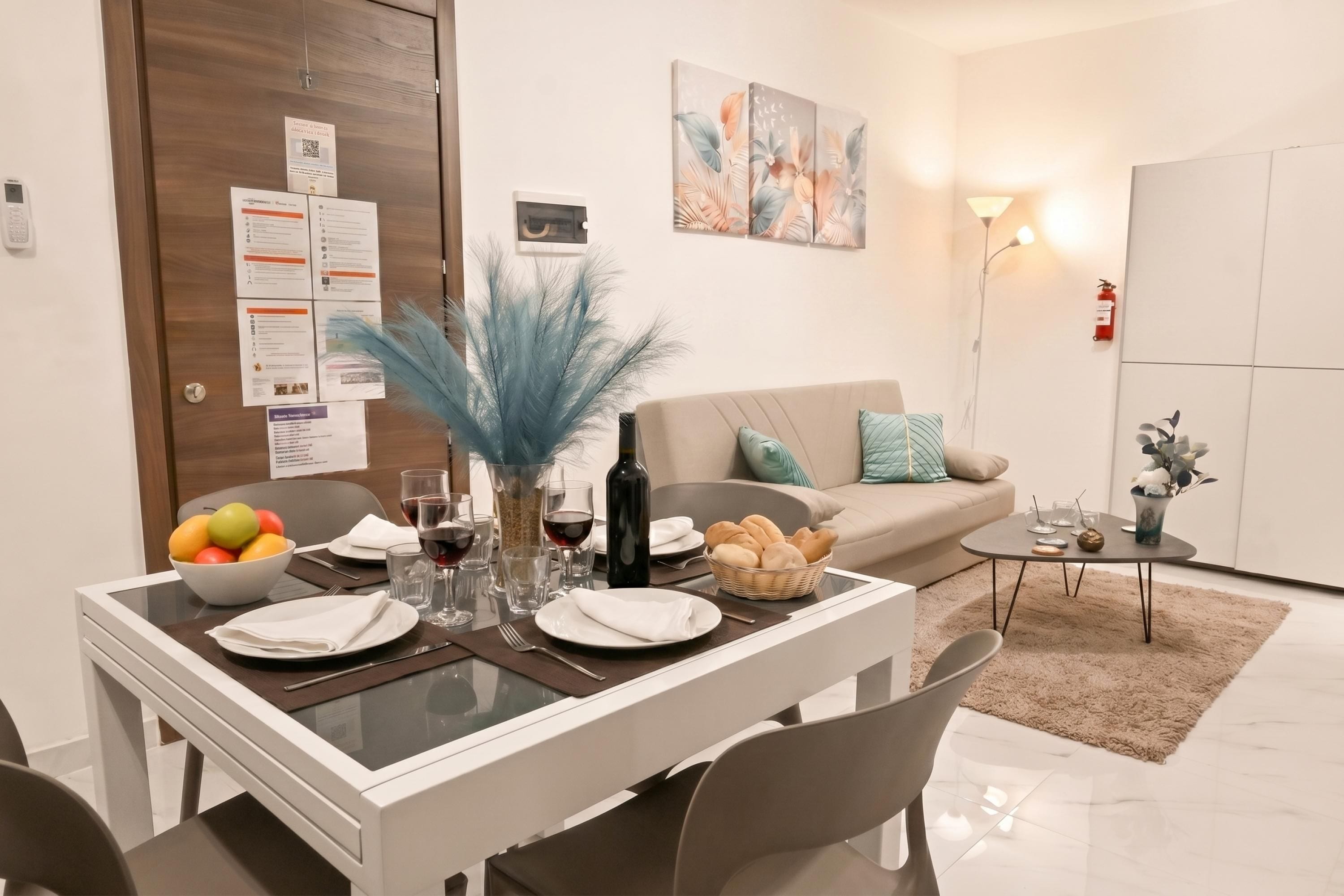 Un comedor con una mesa dispuesta para dos personas, un frutero con fruta, copas de vino y una botella. Se observa un sofá con cojines al fondo, una mesita auxiliar con un florero y un extintor de incendios montado en la pared.