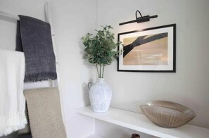 En vit vägg med en inramad abstrakt målning, en vit vas med gröna växter och en beige skål på en vit hylla.