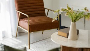 A brown upholstered chair with a metal frame, a round table with a vase of flowers, and a book on the table. - Silkhaus Polo Residence 1BR 2 Baths Meydan (Dubai)