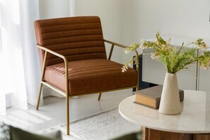 A brown upholstered chair with a metal frame, a round table with a vase of flowers, and a book on the table. - Silkhaus Polo Residence 1BR 2 Baths Meydan (Dubai)