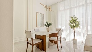 A dining area with a wooden table, four chairs, a vase with a plant, a framed picture on the wall, and sheer curtains. - Silkhaus Polo Residence 1BR 2 Baths Meydan (Dubai)