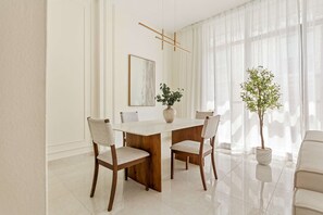 A dining area with a wooden table, four chairs, a vase with a plant, a framed picture on the wall, and sheer curtains. - Silkhaus Polo Residence 1BR 2 Baths Meydan (Dubai)