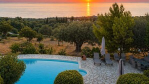 A pool with a stone patio, lounge chairs, and umbrellas, overlooking a sunset over the sea. - Villa Poseidon - Superior Suite In Zante w Pool (Zakynthos)