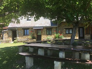 Un bâtiment en pierre avec un toit en pente, des places assises à l’extérieur et un arbre devant.