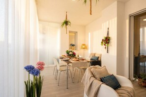 Ein modernes Wohnzimmer mit einer Couch, einem Esstisch und Stühlen. Es gibt ein Fenster mit durchsichtigen Vorhängen, eine hängende Pflanze und eine Lampe auf dem Tisch.
