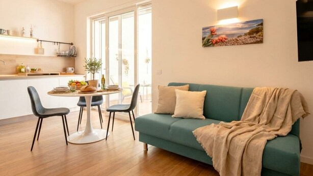 Un salon moderne avec un canapé de couleur turquoise, une table de salle à manger ronde entourée de chaises et un coin cuisine avec une fenêtre.