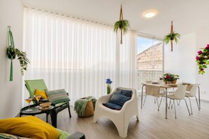 Ein modernes Wohnzimmer mit Essbereich, großen Fenstern mit leichten Vorhängen und hängenden Pflanzen.