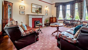A traditional living room with leather sofas, a fireplace, a chandelier, and large windows with floral curtains. - Dunoon - Sleeps 17 - Group Retreat With Parking (Dunoon)