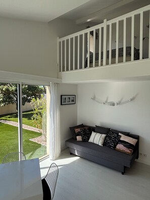 A modern living room with a grey sofa, a dining area with a white table, and a balcony with a railing.