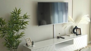 Una sala de estar moderna con una televisión de pantalla plana montada en la pared, un mueble de entretenimiento blanco con un marco de fotos, una planta en maceta y una copa de vino sobre la mesa.