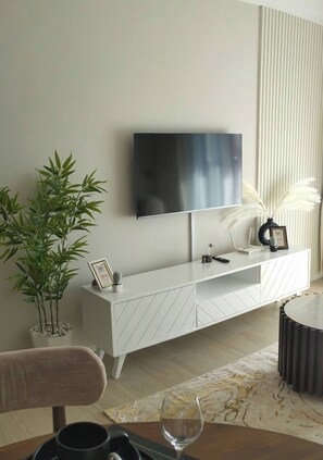 Ein modernes Wohnzimmer mit einem flachbildfernseher an der Wand, einer weißen Unterhaltungsanlage mit Bilderrahmen, einer Topfpflanze und einem Glas Wein auf dem Tisch.