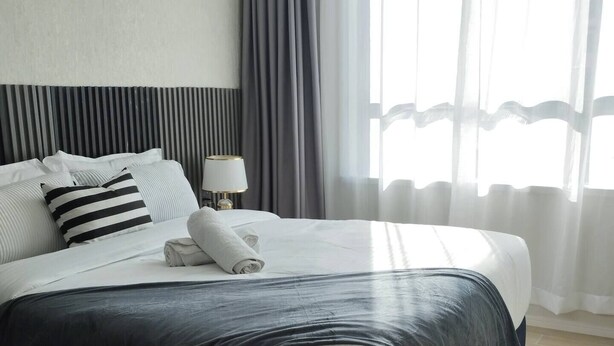 Una cama bien hecha con un cabecero oscuro, una lámpara de noche y una ventana con cortinas traslúcidas.