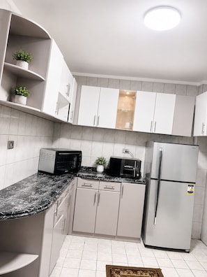 A modern kitchen with white cabinets, a black countertop, a microwave, a toaster, a refrigerator, and a small potted plant. - Spacious Apartment Cairo (Cairo Governorate)