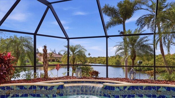 A pool with a waterfall feature, surrounded by tropical plants and palm trees, with a view of a lake and houses in the background. - Mar3507 3 Bedroom Home (Punta Gorda)
