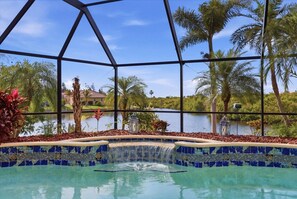 A pool with a waterfall feature, surrounded by tropical plants and palm trees, with a view of a lake and houses in the background. - Mar3507 3 Bedroom Home (Punta Gorda)