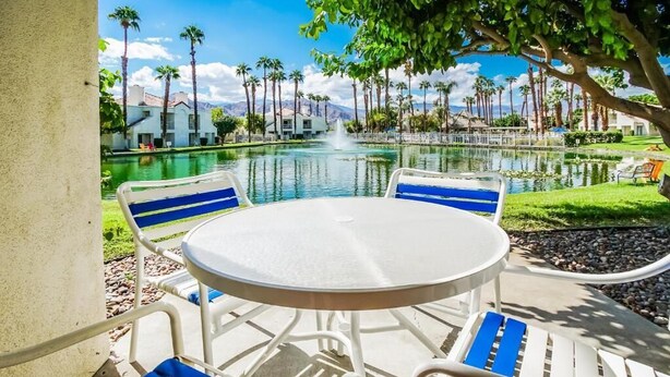 Un espace aménagé au bord de la piscine, avec une table ronde et des chaises, une fontaine, et une vue sur les palmiers et les bâtiments.