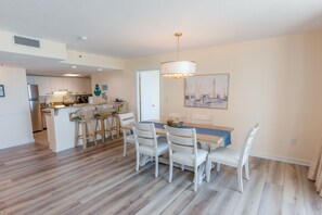 A dining area with a table and chairs, a kitchen in the background, and a painting on the wall. - 329 North Hampton, Expansive Balcony & Resort Amenities 2 Bedroom Condo (Myrtle Beach)