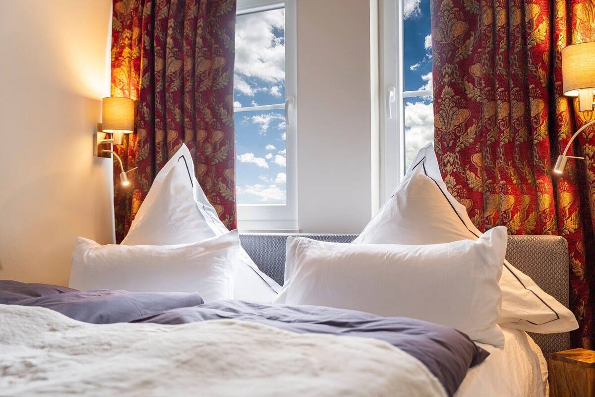 A hotel room with a bed, two white pillows, a window with a view of the sky, and red patterned curtains.