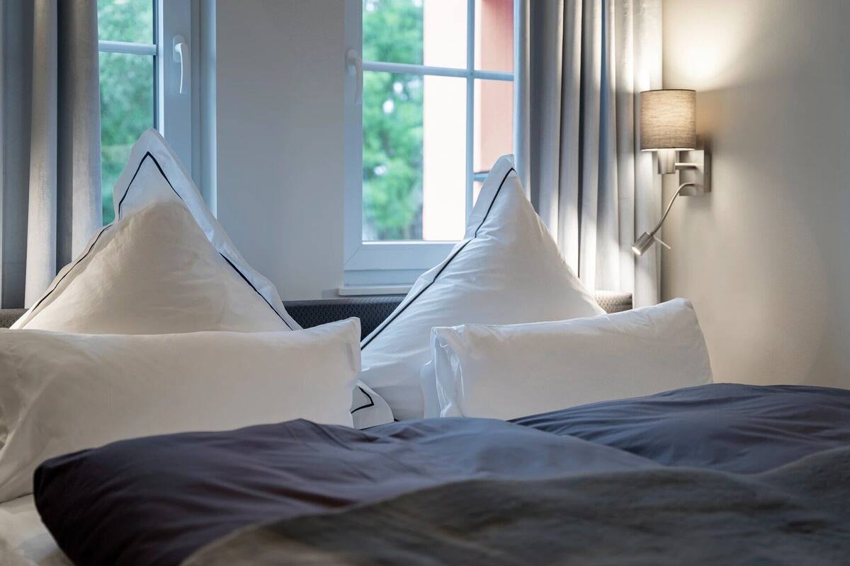 A hotel room with a bed, white pillows, a window with curtains, and a wall-mounted lamp.