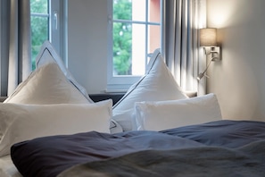 Ein Hotelzimmer mit Bett, weißen Kissen, einem Fenster mit Vorhängen und einer Wandlampe.